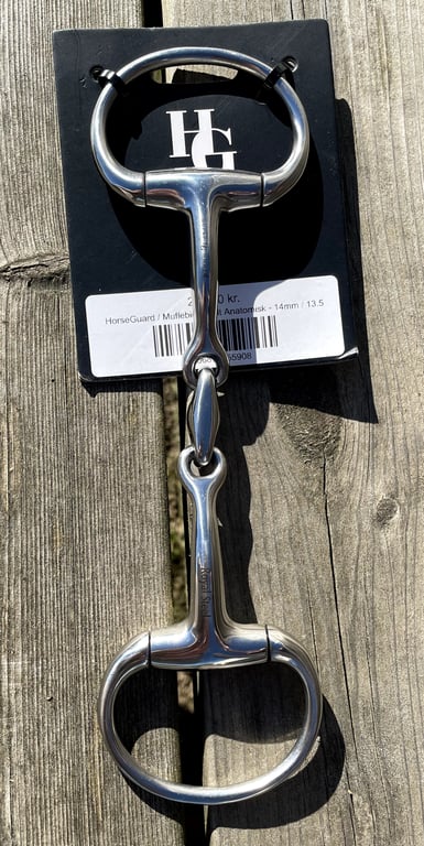 Biddestykke i rustfrit stål med to ringe og leddelt bid, lagt på en træflade med en sort baggrundslabel med logoet "HG".