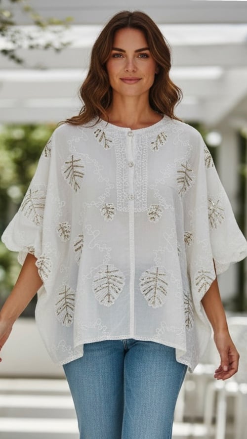 Kvinde iført hvid, løs bluse med broderede bladmotiver og blå jeans, stående i lyst, moderne rum. (1)