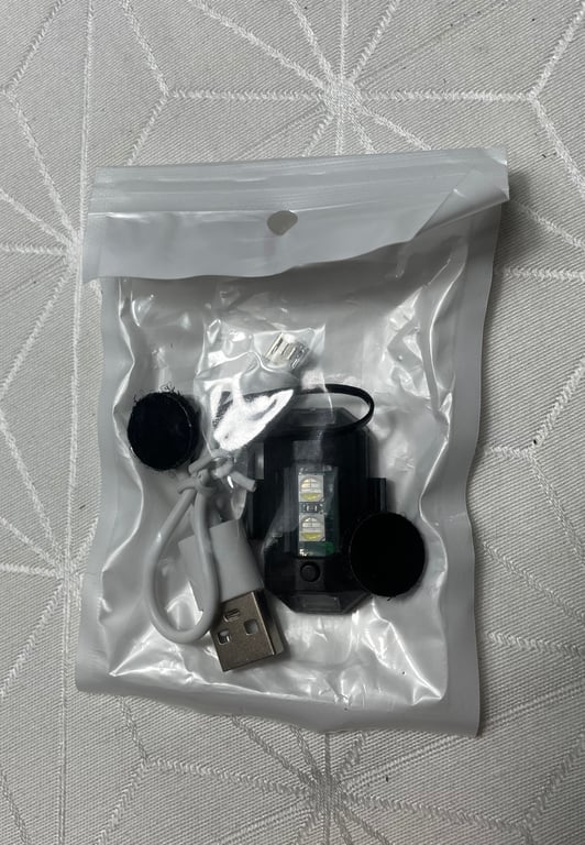 Plastpose med USB-kabel og lille elektronisk enhed med LED-lys, placeret på et tekstureret, lyst underlag.