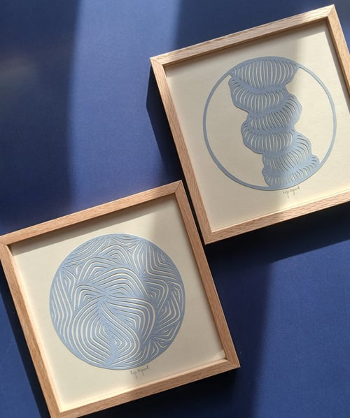 To indrammede kunsttryk med abstrakte blå cirkel- og bølgemønstre på beige baggrund, placeret på blå overflade med sollys. (2)