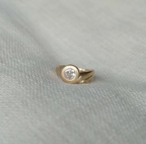 Guld ring med rund diamant sat i midten, placeret på lyst tekstileret stof. (1)