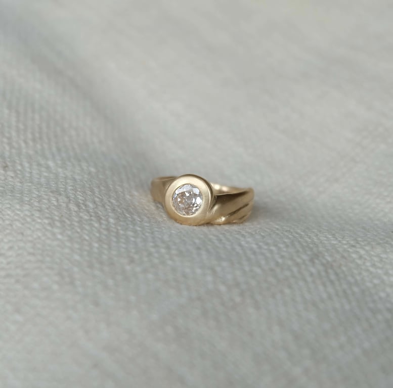 Guld ring med rund diamant sat i midten, placeret på lyst tekstileret stof.