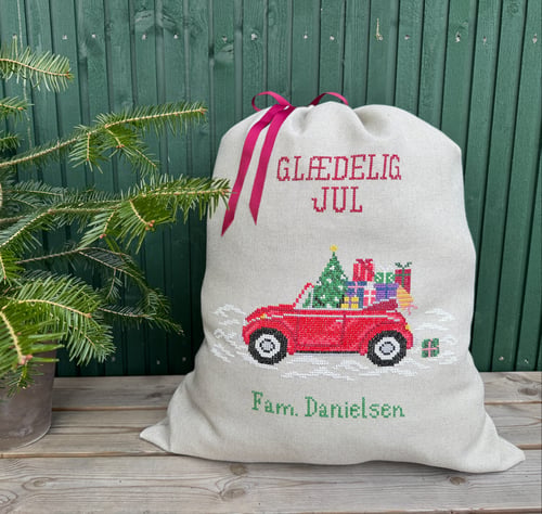 Julesæk med broderet rød bil fuld af gaver, teksten "GLÆDELIG JUL" og "Fam. Danielsen," placeret ved siden af en lille juletræ i potte foran en grøn trævæg. (1)
