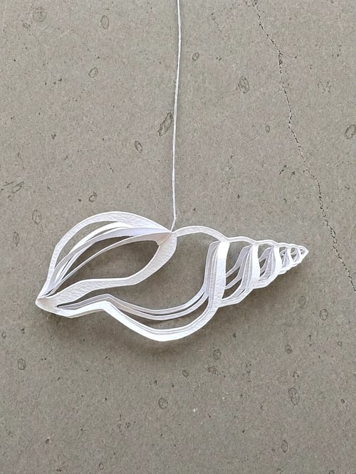 Hvid papirskulptur formet som en konkylie hængende i en snor mod en grå baggrund. (1)