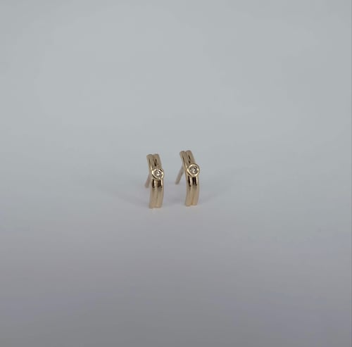 Små øreringe i guld med en enkelt rund diamant i midten, vist på grå baggrund. (1)