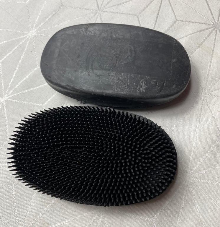 To sorte hårbørster i oval form, den ene med børstehår og den anden med glat overflade, placeret på en lysegrå dug med geometrisk mønster.