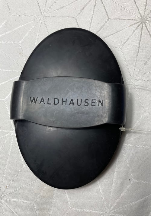 Sort oval ruskindsbørste med rem og teksten "WALDHAUSEN" på, lagt på hvidt mønstret stof. (1)