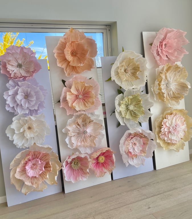 Store papirblomster i pastelfarver monteret på hvide plader, opstillet mod en væg med et vindue i baggrunden.