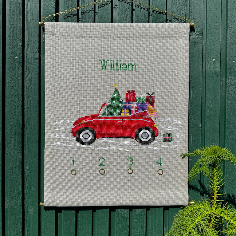 Broderet kalenderspænd med en rød bil fyldt med julegaver og et juletræ, navnet William øverst og tal fra 1 til 4 nederst med små metalringe.
