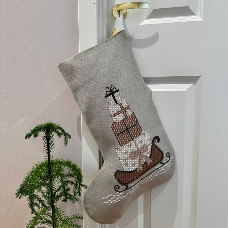 Julesok i gråt stof med broderi af sække med gaver på en slæde hængende på en dørhåndtag, ved siden af en lille grøn plante.