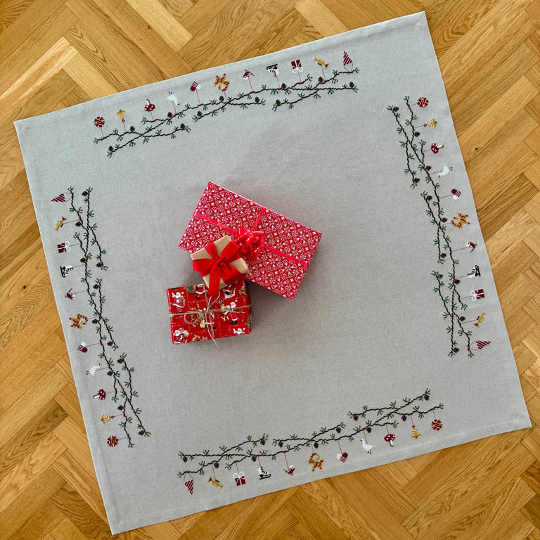 Et gråt juledug med broderede julemotiver langs kanten og to indpakkede julegaver med rødt gavepapir og sløjfer placeret midt på dugen på et trægulv.