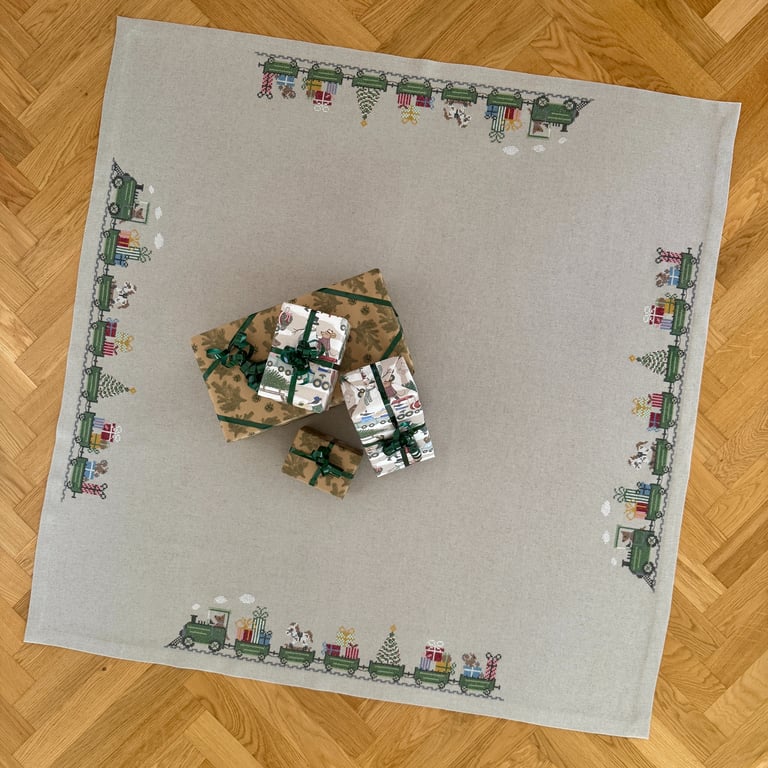Gråt juledug med broderet tog med gaver, juletræer og dyr langs kanten, fire indpakkede gaver med bånd midt på dugen, lagt på trægulv.
