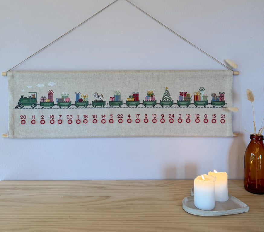 Broderet julekalender med et grønt tog pyntet med julegaver og en juletræ på en hørstof, hængende på en væg over et lyst træbord med to tændte hvide stearinlys og en brun glasvase med tørret græs.