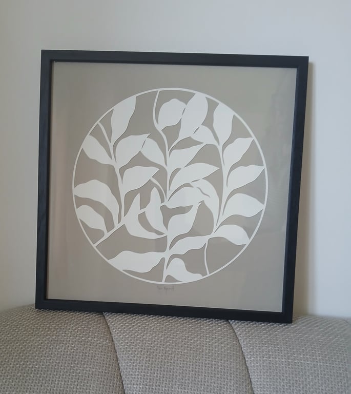 Sort ramme med minimalistisk planteillustration i hvid på beige baggrund placeret på en tekstureret lysegrå sofa.