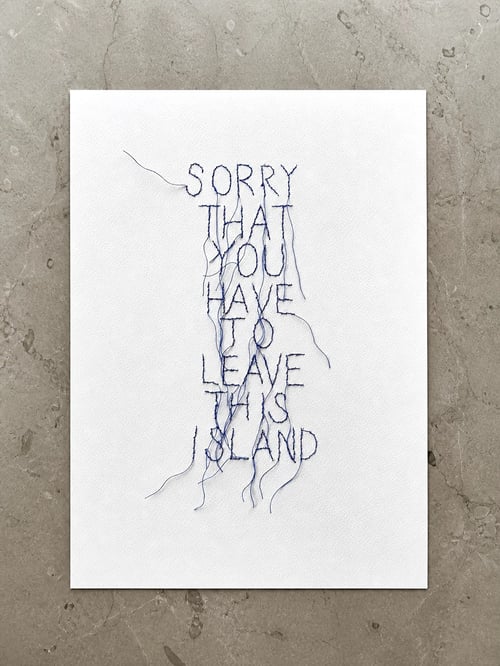 Hvidt stykke stof med håndbroderet tekst i blå tråd: "SORRY THAT YOU HAVE TO LEAVE THIS ISLAND". Tråde hænger løst ned fra bogstaverne. Stof ligger på grå marmorbaggrund. (1)