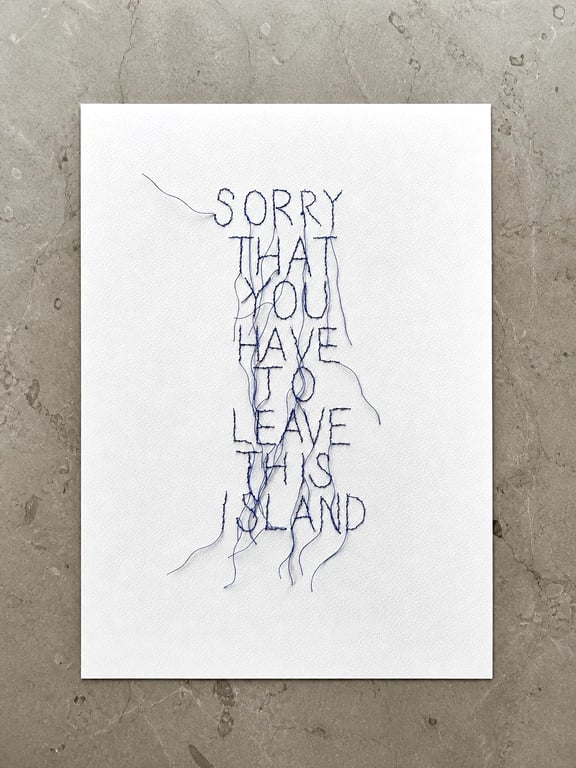 Hvidt stykke stof med håndbroderet tekst i blå tråd: "SORRY THAT YOU HAVE TO LEAVE THIS ISLAND". Tråde hænger løst ned fra bogstaverne. Stof ligger på grå marmorbaggrund.