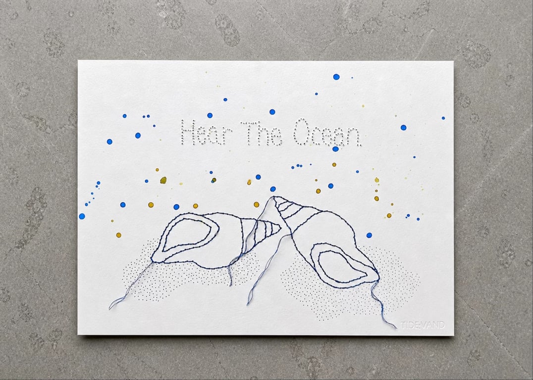 Et hvidt lærred med broderi af to blå muslinger og teksten "Hear The Ocean" øverst. Der er blå og gule farvepletter spredt over lærredet, og nederst til højre står ordet "TIDEVAND".