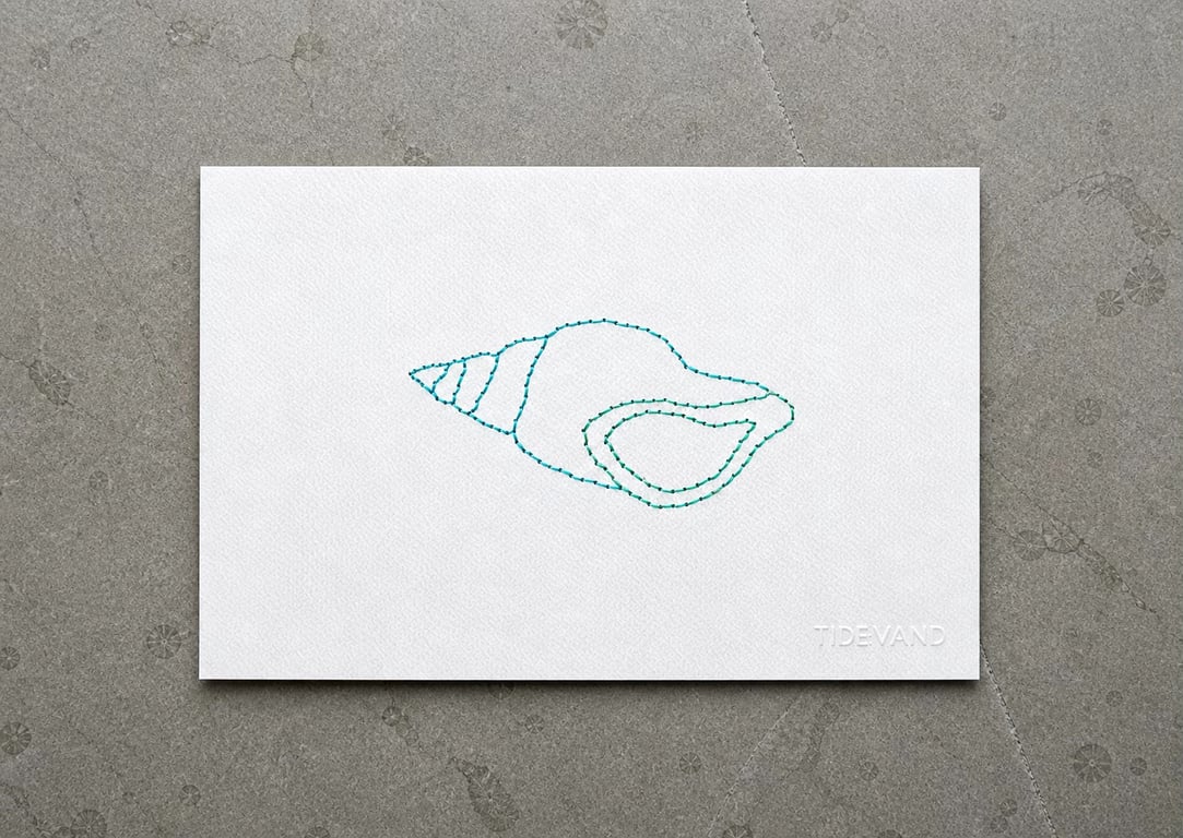 Hvidt papir med broderet, enkelt kontur af en snegleskal i blå og grønne tråde, lagt på en grå stenoverflade.