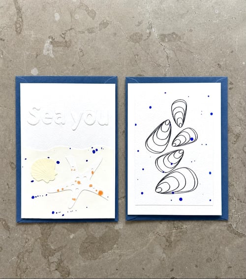 To minimalistiske kunstkort med blå kanter, den ene med et sømandsmotiv med teksten "Sea you" og en søstjerne, den anden med sort-hvide muslingeskaller og blå prikker, begge på en grå betonoverflade. (1)