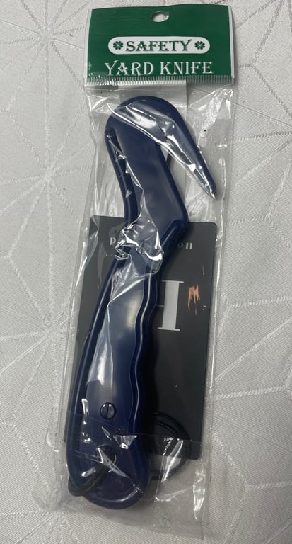 Sikkerhedsmesser i blå plastik, indpakket i gennemsigtigt plast med grøn og hvid etikette med teksten "SAFETY YARD KNIFE".