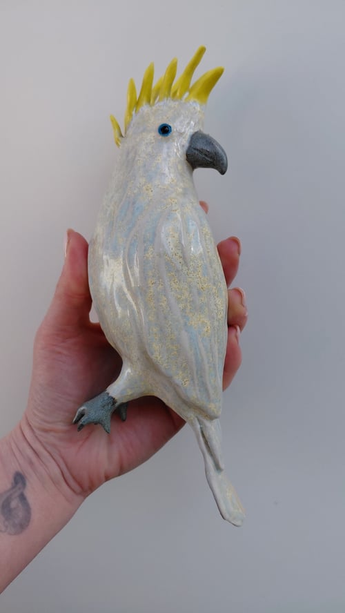 Hånd holder en keramisk figur af en hvid kakadue med gul kam og blå øjne mod en lys baggrund. (1)
