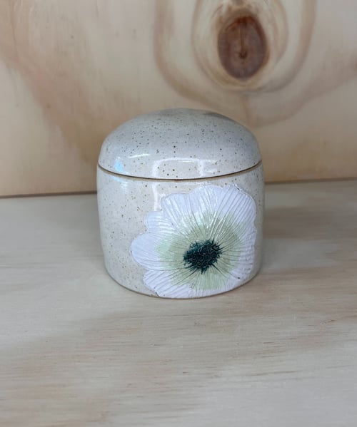 Lille, rund keramikbeholder med låg, dekoreret med et malet hvidt og grønt blomstermotiv, placeret på et lyst træbord mod en baggrund af lyst træ. (1)