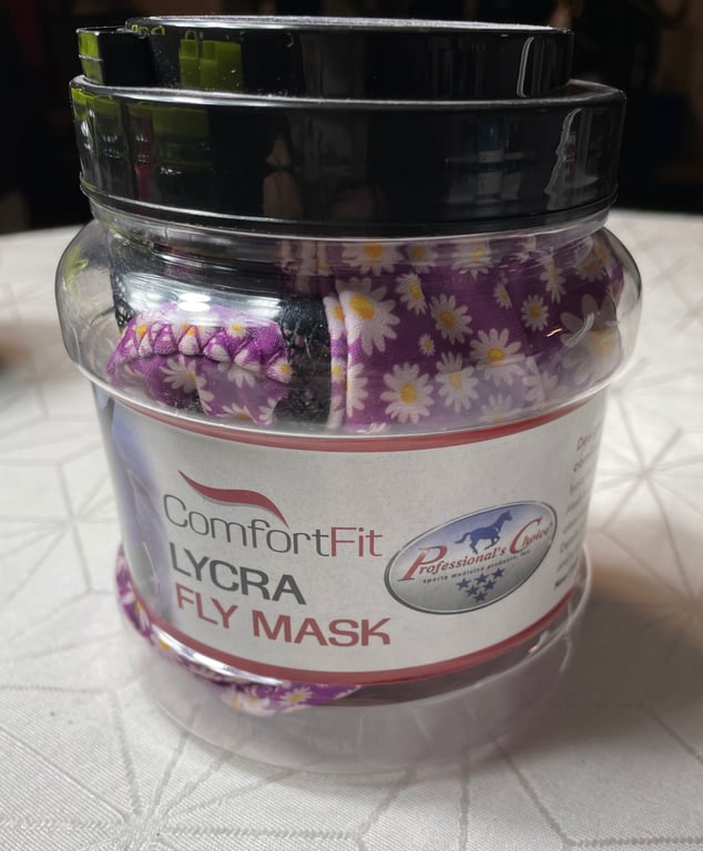 Transparent plastbeholder med sort låg indeholdende en lilla flyvemaske med hvid-gule blomster, mærket "ComfortFit Lycra Fly Mask" fra Professional's Choice.