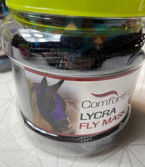 En gennemsigtig plastikbeholder med en limegrøn låg, der indeholder en netpose og en etiket med et billede af en hest iført en lilla Lycra fluenetmaske og teksten "ComfortFit LYCRA FLY MASK". (1)