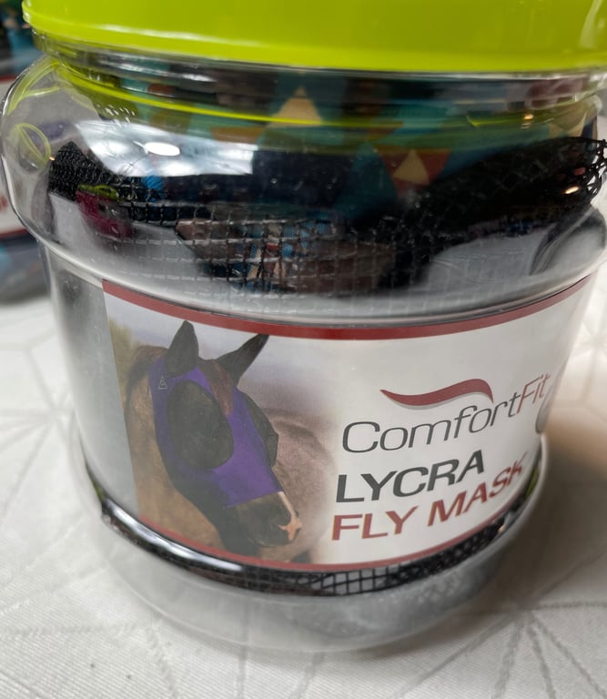 En gennemsigtig plastikbeholder med en limegrøn låg, der indeholder en netpose og en etiket med et billede af en hest iført en lilla Lycra fluenetmaske og teksten "ComfortFit LYCRA FLY MASK".