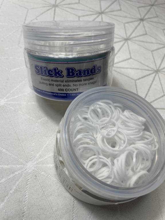 To behold a treasure trove of hairstyling convenience in a crisp, angled view of two transparent plastic containers filled with white elastic hair bands labeled "Slick Bands," resting on a textured, patterned white surface, evoking neatness and ease.