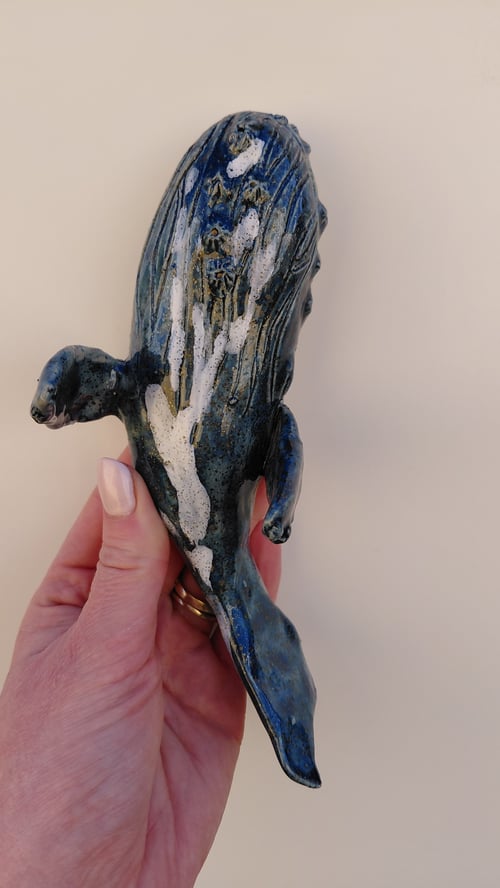Hånd, der holder en lille keramisk blåhval med hvide detaljer mod en lys baggrund. (1)