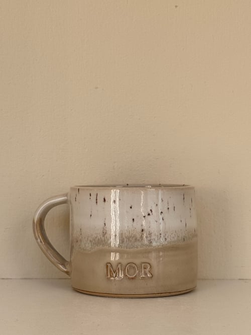 Keramikkrus med glaseret overflade i beige og hvid, hvor ordet "MOR" er præget på forsiden. (1)