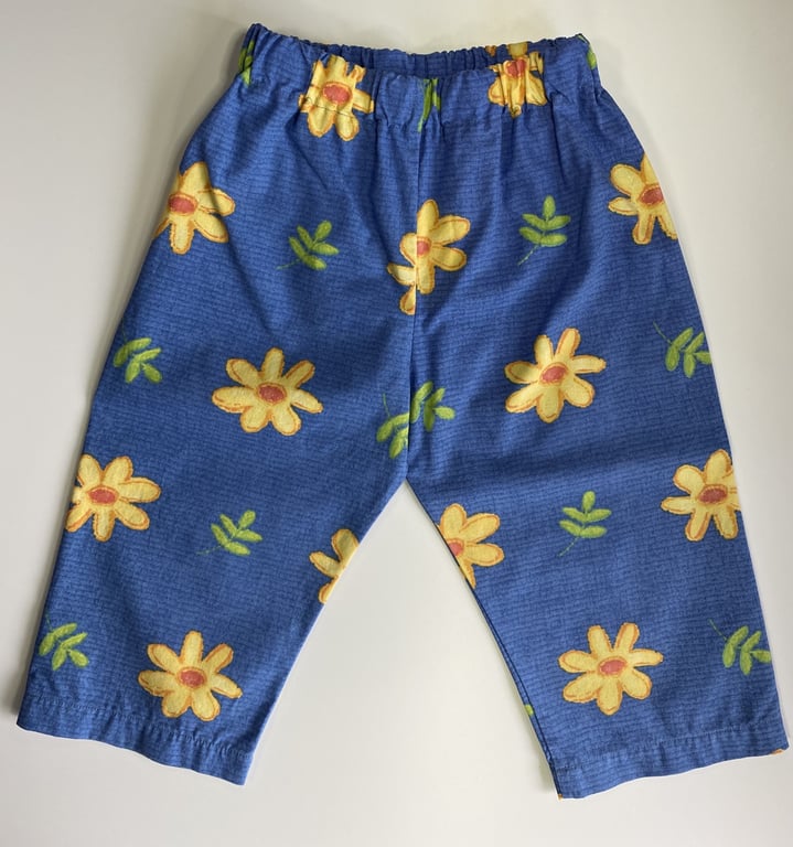 Blå børnebukser med elastisk talje og mønster af store gule blomster og grønne blade.