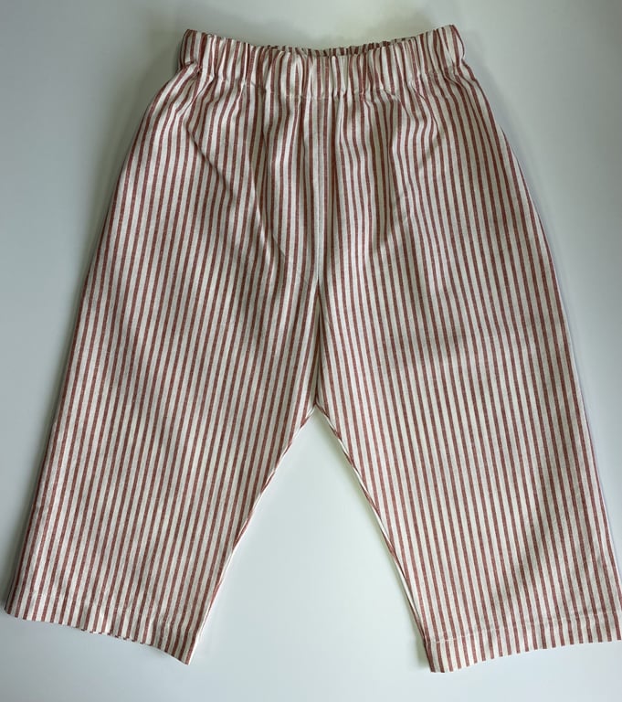 Bukser til baby eller små børn med elastisk talje og smalle røde og hvide lodrette striber, lagt fladt på hvid baggrund.