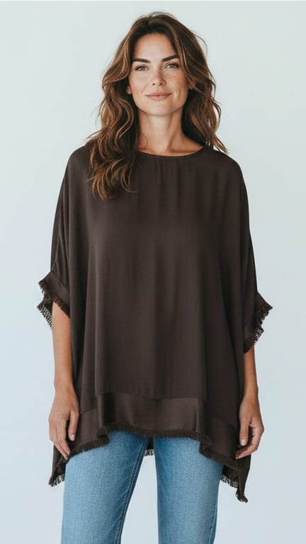 Kvinde iført oversized mørkebrun bluse med frynser og lyseblå jeans, stående mod en lyseblå baggrund.