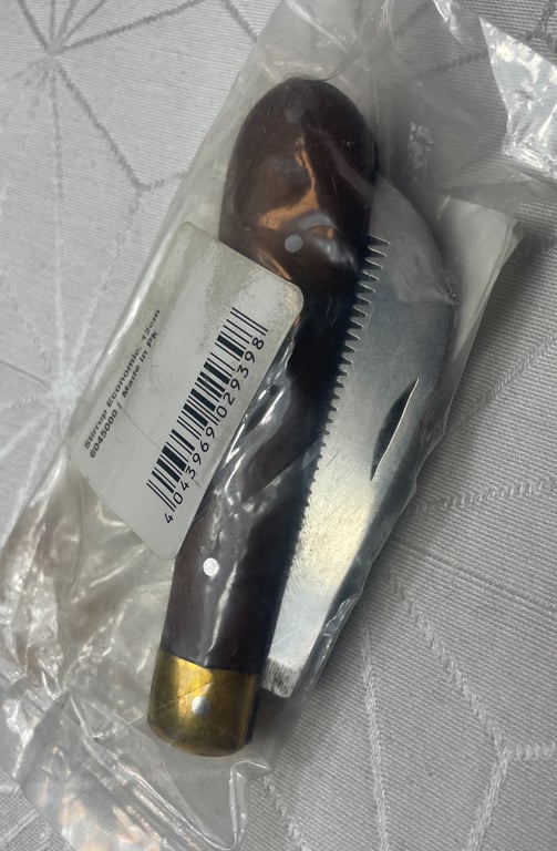 Kniv med tandet blad og træskaft i plastpose med pris-etiket, lagt på stof med geometrisk mønster.