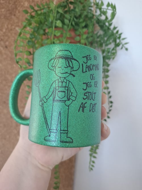 Grøn glitrende krus med tegning af en landmand og teksten "Jeg er landmand og jeg er stolt af det", holdt i en hånd med en grøn plante i baggrunden. (1)