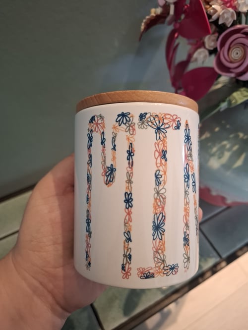 Hånd, der holder en hvid krukke med bamboo låg og blomsterformede mønstre i blå, orange og grå. (1)