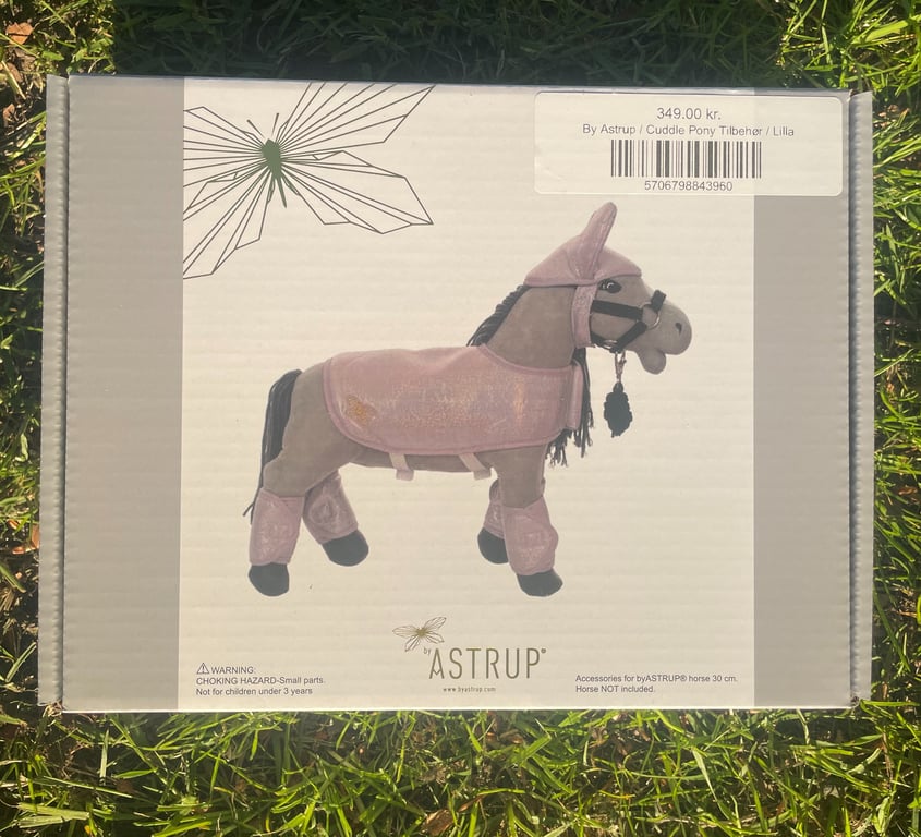 Æske med by Astrup Cuddle Pony tilbehør i lilla, vist på græs.
