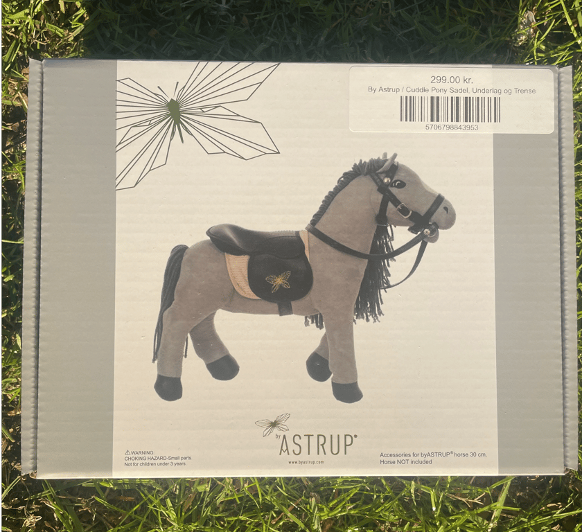 Pakke med tilbehør til byASTRUP Cuddle Pony, inklusive sadel, underlag og trense, på græs.