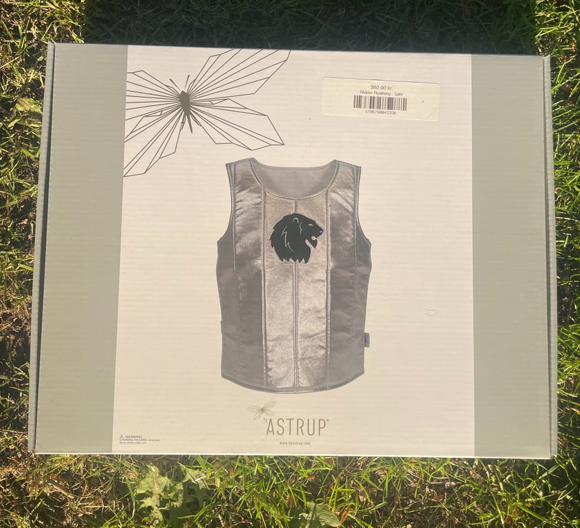 Æske med billede af sølvfarvet riddervest med løve-logo, mærket "Astrup", placeret på græs.