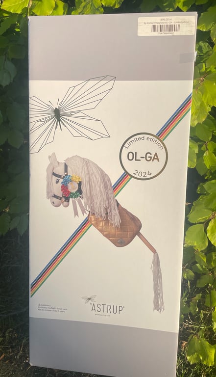 Æske med billedet af en limited edition hobbyhest med blomsterdetaljer og teksten "OL-GA 2024" fra Astrup, stående mod grønne blade.
