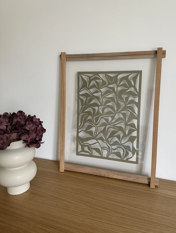Et glasbillede med abstrakte grønne blade i en træramme, placeret på et lyst træbord ved siden af en hvid vase med mørkelilla blomster.