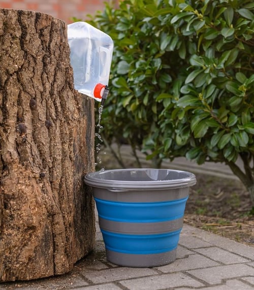 En plastflaske hældt op mod en træstub med vand, der drypper ned i en foldbar blå og grå spand på stenbelagt jord med grønne buske i baggrunden. (1)