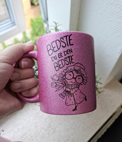 En hånd holder en glitrende pink kop med teksten "BEDSTE DU ER DEN BEDSTE" og en tegning af en glad person med briller, hat og et tørklæde, der holder en buket blomster. (2)