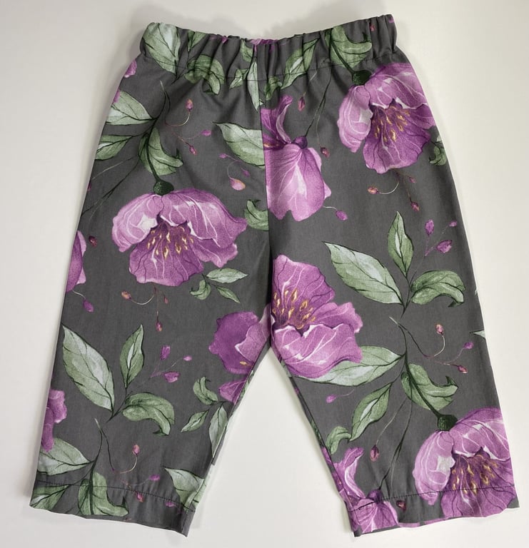 Bukser til børn med elastisk talje i grå stof med print af store lilla blomster og grønne blade.