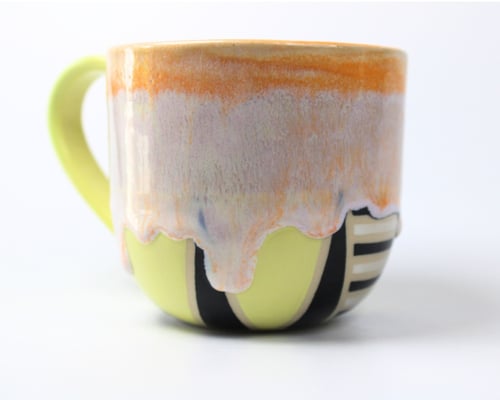En keramisk kop med håndtag, dekoreret med orange, lilla og lysegrøn glasur, der drypper ned over en sort og beige geometrisk mønster. (1)