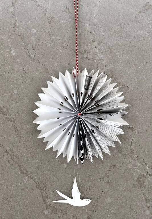 Origami-figur lavet af foldet papir med tekst, ophængt mod en grå væg med en rød-hvid snor og en hvid fuglefigur hængende under.