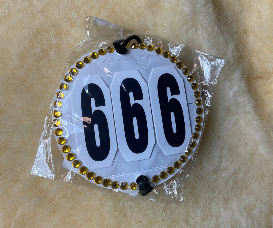 Rundt, hvidt mærke med tallet 666 i sort, dekoreret med gule perler langs kanten og pakket i klar plastik.