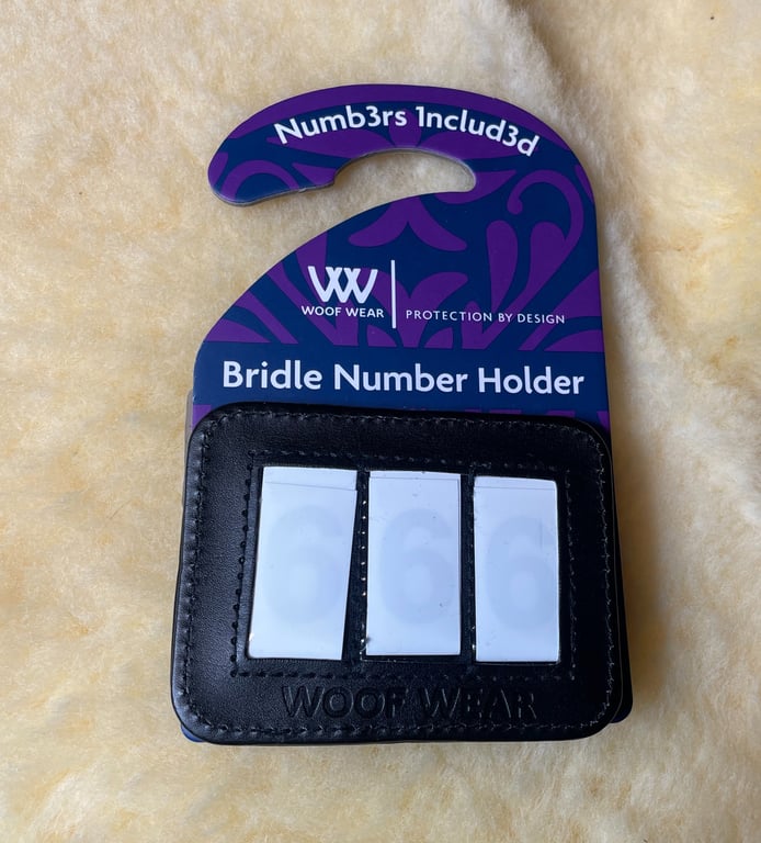Sort læderholder til nummer med tre hvide indstillelige tal, mærket "Woof Wear", på lilla og blå emballage med teksten "Bridle Number Holder" og "Numb3rs 1nclud3d".
